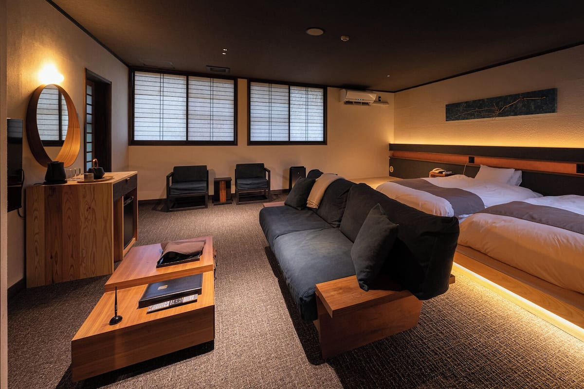 A hotel room with two beds, a sofa, a small desk, a mirror, and a window with blinds.