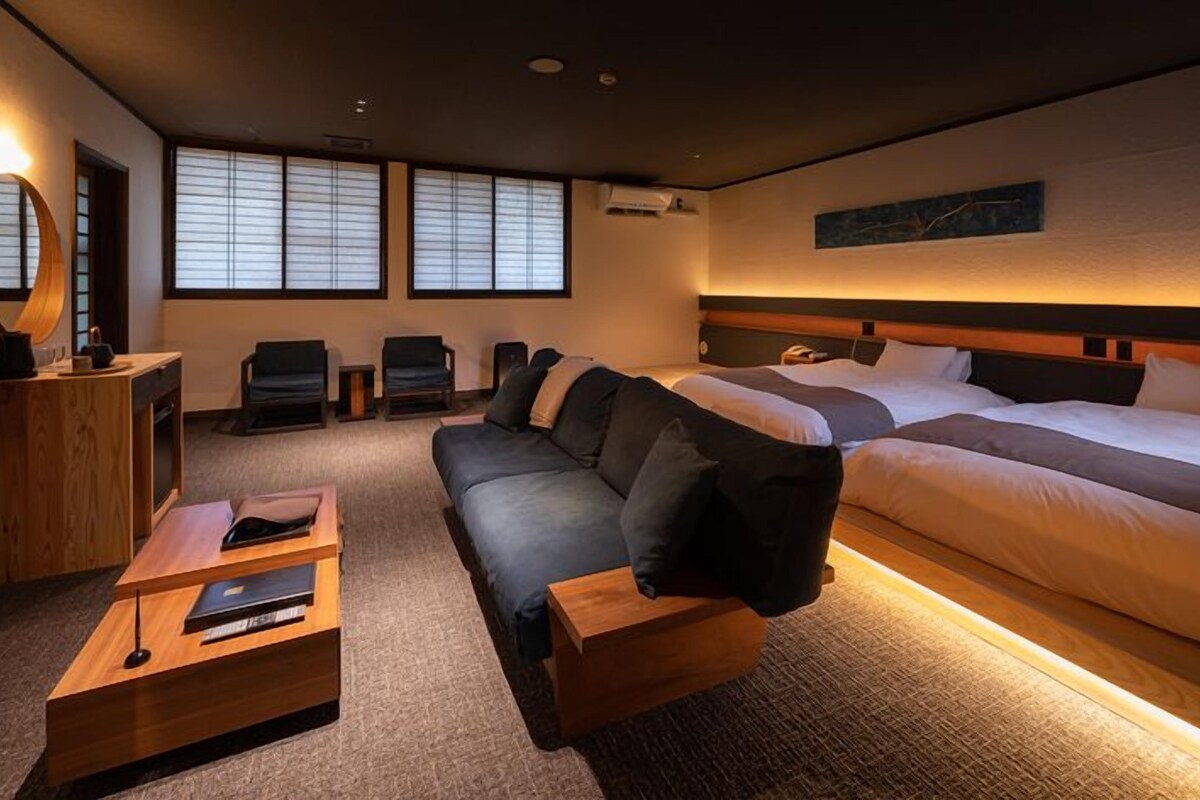 Une chambre d’hôtel avec deux lits, un canapé, une table d’appoint en bois, un miroir et une fenêtre avec des stores.