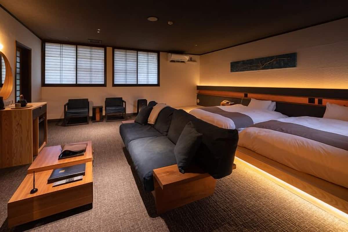A hotel room with two beds, a sofa, a wooden side table, a mirror, and a window with blinds.