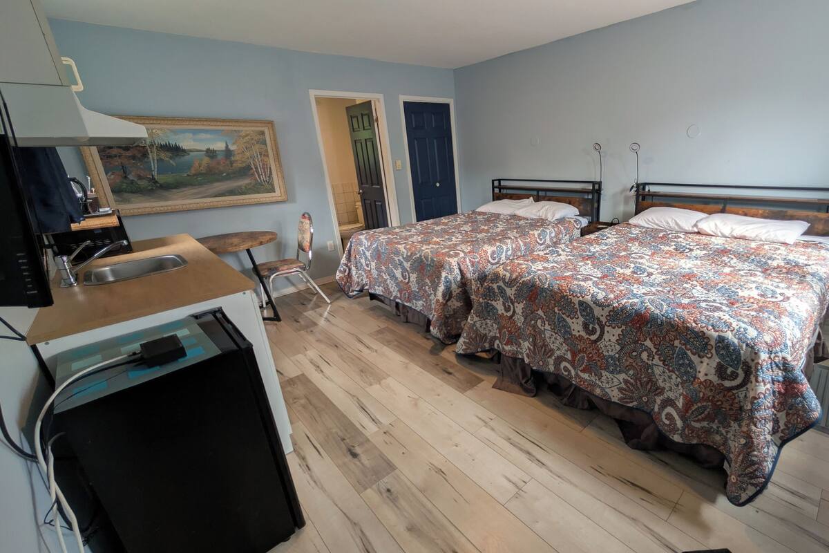 A bedroom with two beds, a wooden desk with a chair, a painting on the wall, and a door leading to another room.
