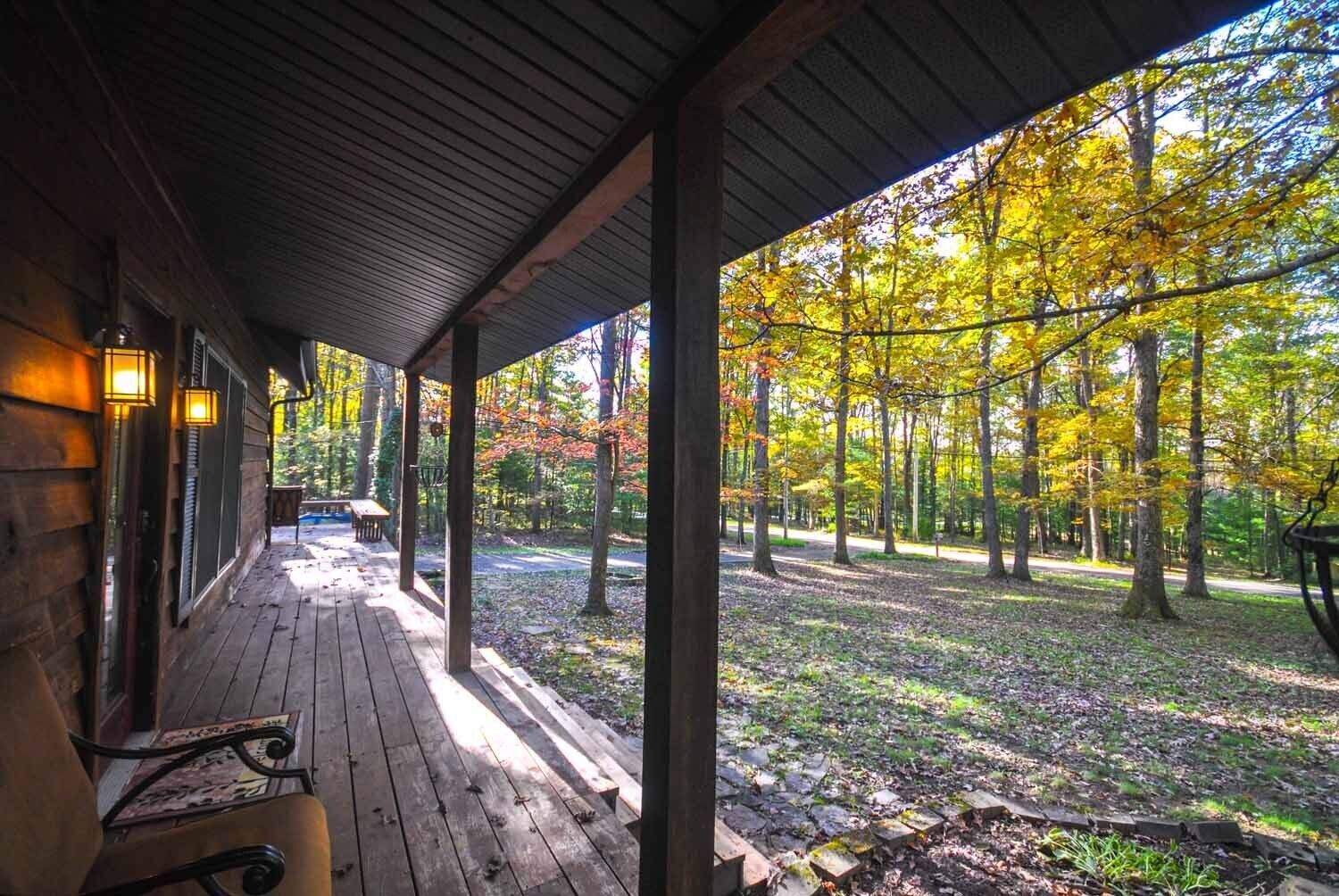 Un porche de madera con un banco y una mesa, con vistas a un bosque de follaje otoñal.