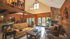 A spacious living room with a high ceiling, wooden beams, a fireplace, and a large window. - Corey Cabin - Cozy Getaway Minutes from Ohiopyle (Ohiopyle)