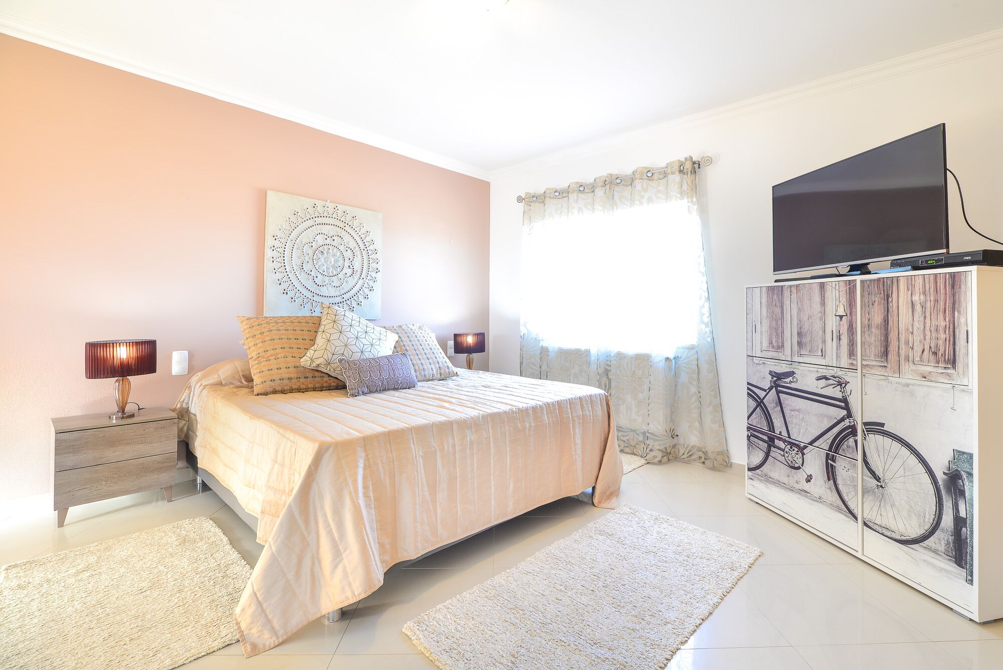 Une chambre à coucher comprenant un lit, des lampes de chevet, une œuvre d’art murale, une fenêtre avec des rideaux et un téléviseur fixé au mur.