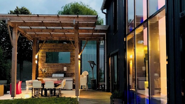 Un espace extérieur moderne, aménagé en terrasse, avec une pergola en bois, des sièges et une pièce fermée par des parois de verre.