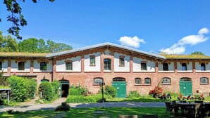 Un edificio grande de ladrillo con múltiples ventanas y puertas, rodeado de abundante follaje y mobiliario para el exterior.