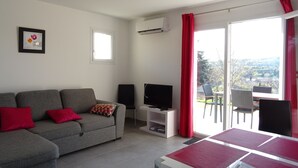 Een moderne woonkamer met een grijze bank, rode kussens, een flatscreen televisie en een eettafel met een rode tafelkleed.