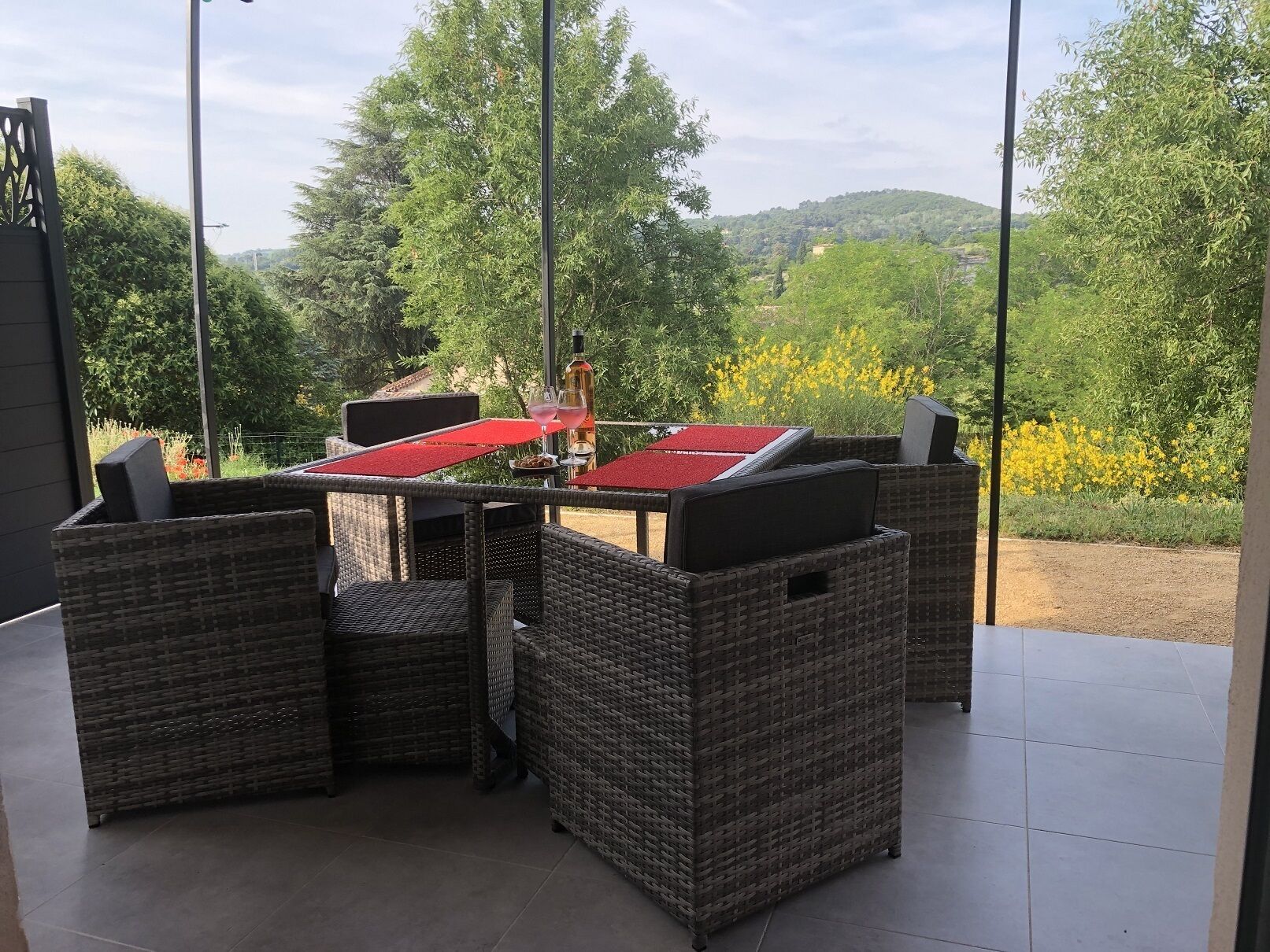 An outdoor patio with wicker furniture and a table set for dining, overlooking a scenic landscape with trees and hills.