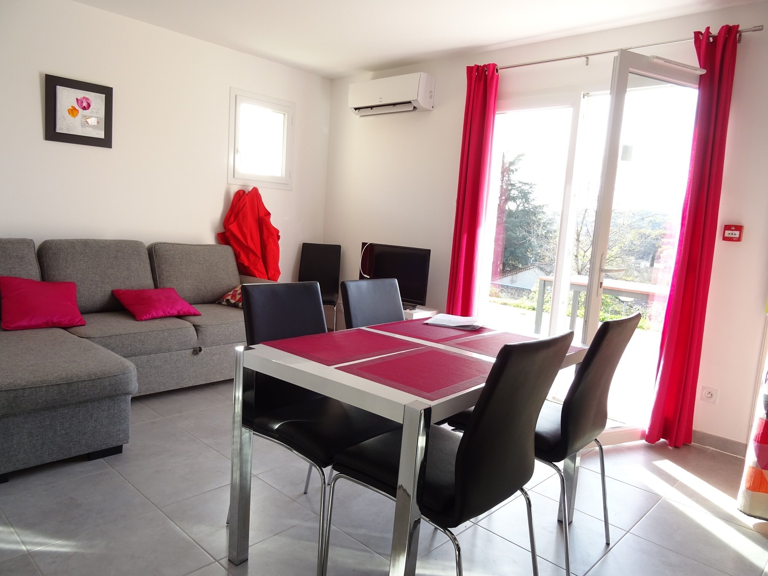 Ein Wohnzimmer mit grauem Sofa, roten Kissen, einem Esstisch mit Stühlen und einem roten Vorhang.