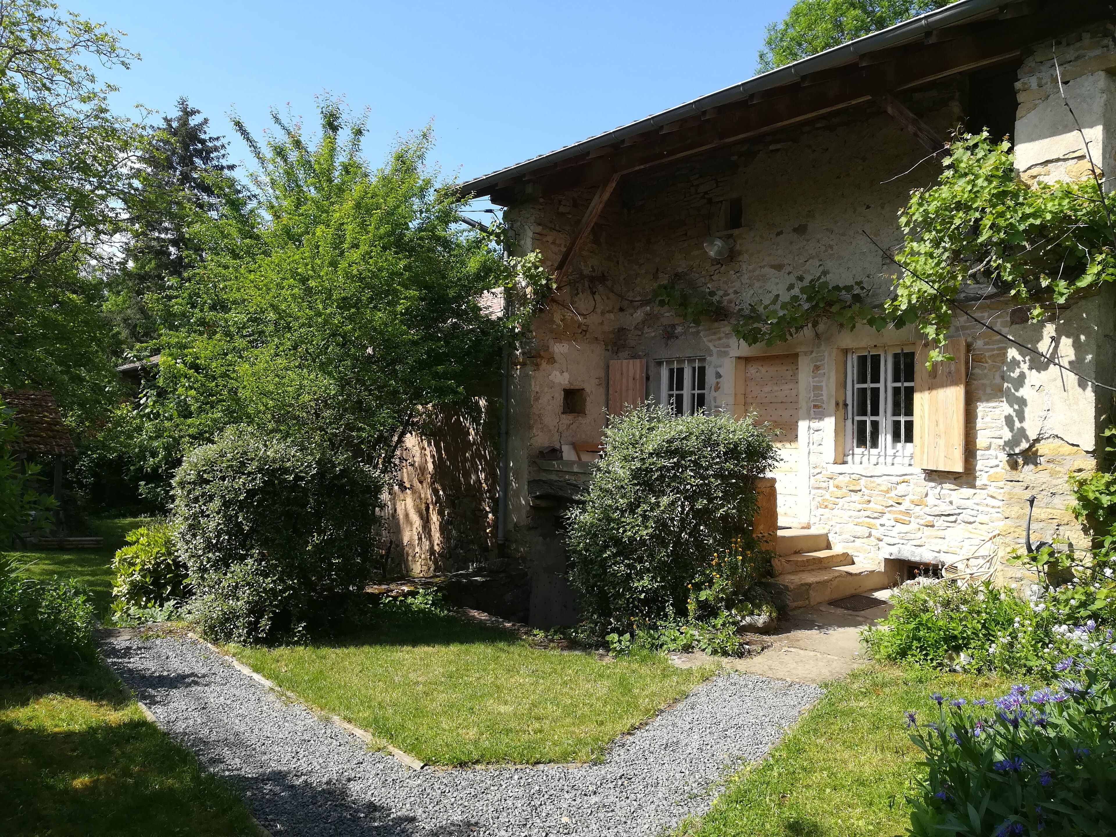 A rustic stone house with a gravel pathway leading to the entrance, surrounded by lush greenery and flowering plants.