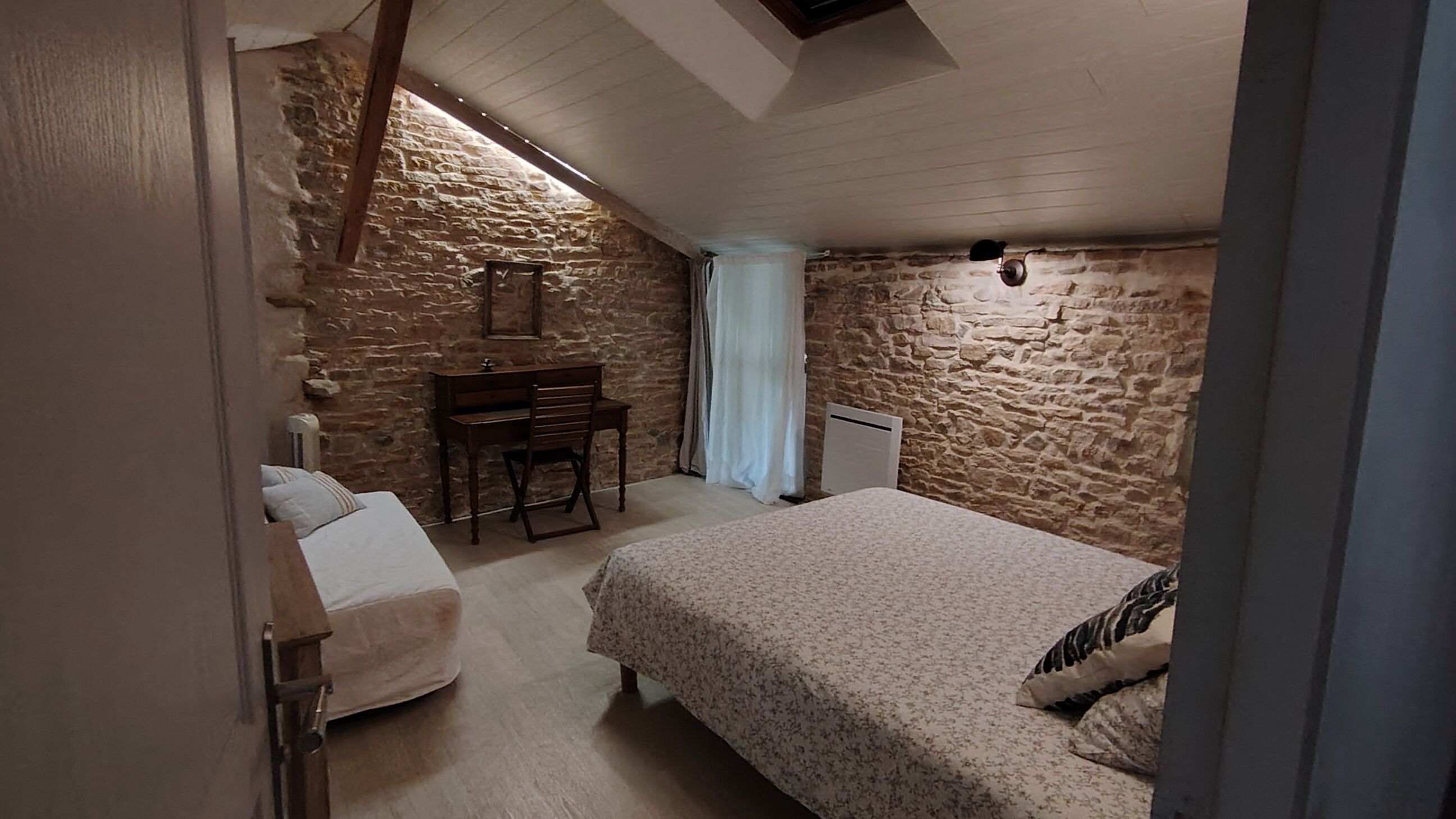 Een slaapkamer met een stenen muur, een bed, een bureau met stoel en een radiator.