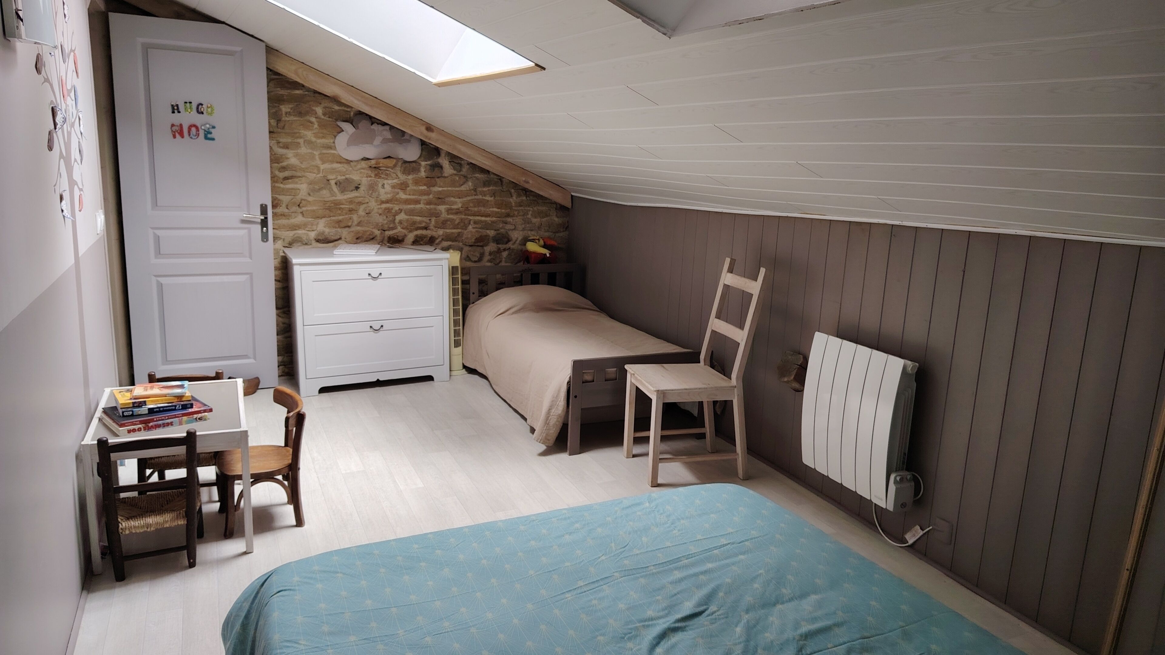 A bedroom with a loft bed, a desk with books, a chair, a radiator, and a stone wall.