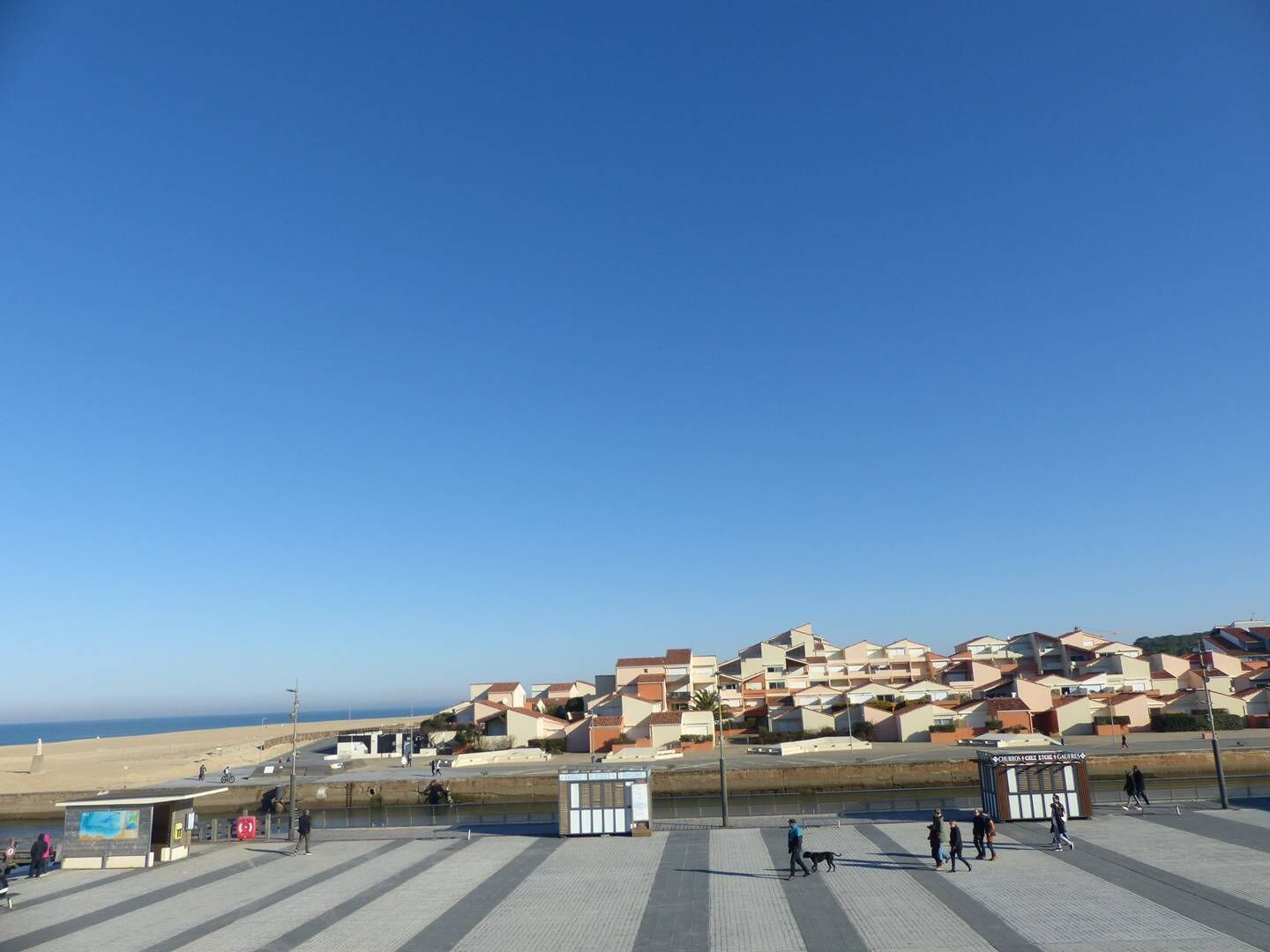A coastal promenade with a tiled walkway, a small kiosk, and residential buildings in the background.