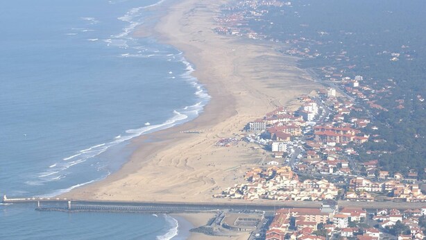 Vista aérea de un pueblo costero con playa, muelle y un conjunto denso de edificios con tejados rojos.