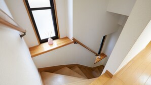 Un escalier en bois avec une rampe, une petite étagère sur laquelle est posé un vase rose, et une fenêtre laissant entrer la lumière naturelle.