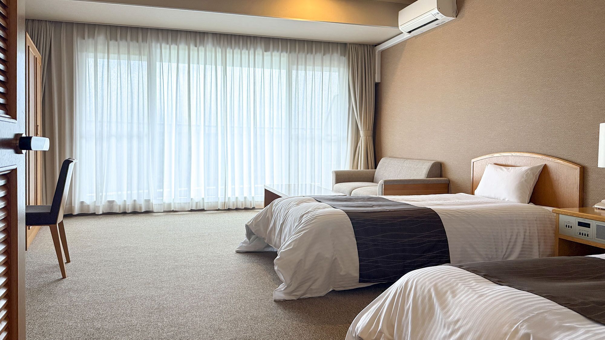 Habitación de hotel con dos camas, un escritorio, una silla y un ventanal con cortinas traslúcidas.