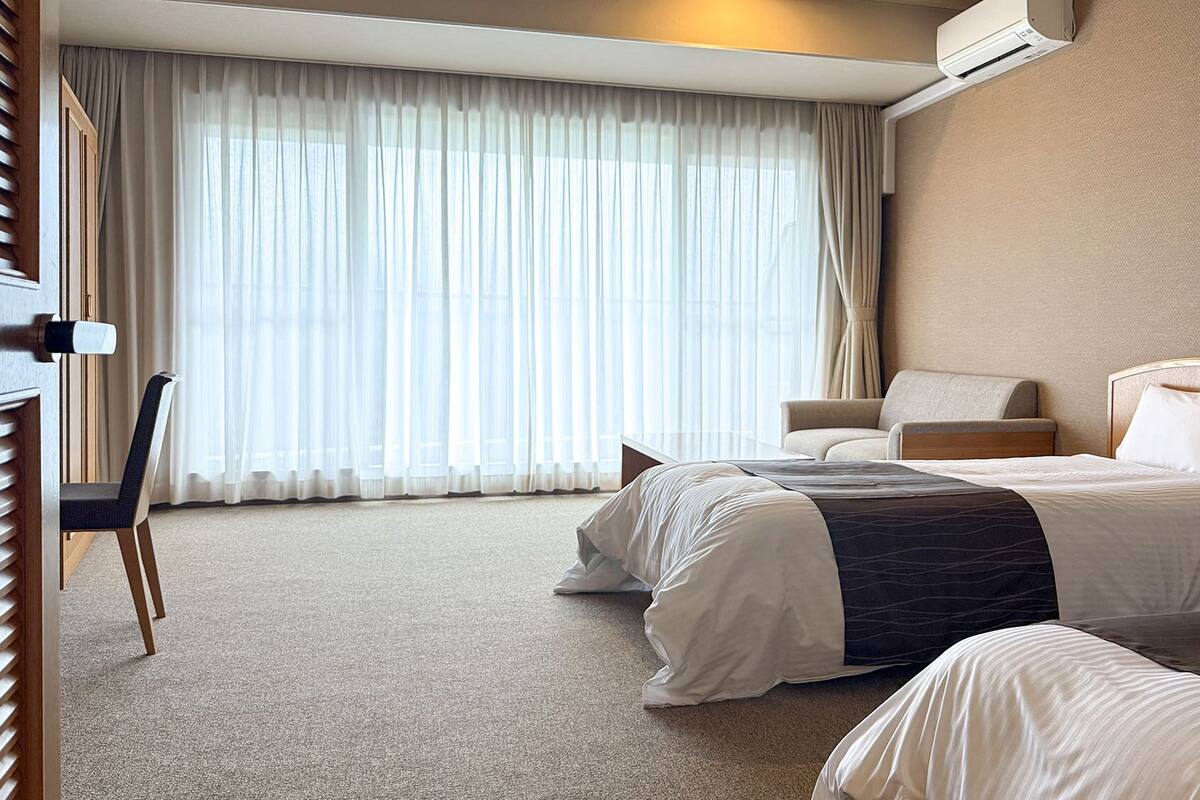 Habitación de hotel con dos camas, un escritorio, una silla y un ventanal con cortinas traslúcidas.