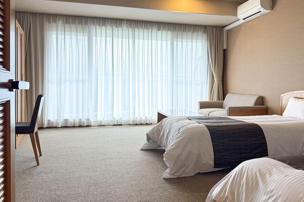 Habitación de hotel con dos camas, un escritorio, una silla y un ventanal con cortinas traslúcidas.