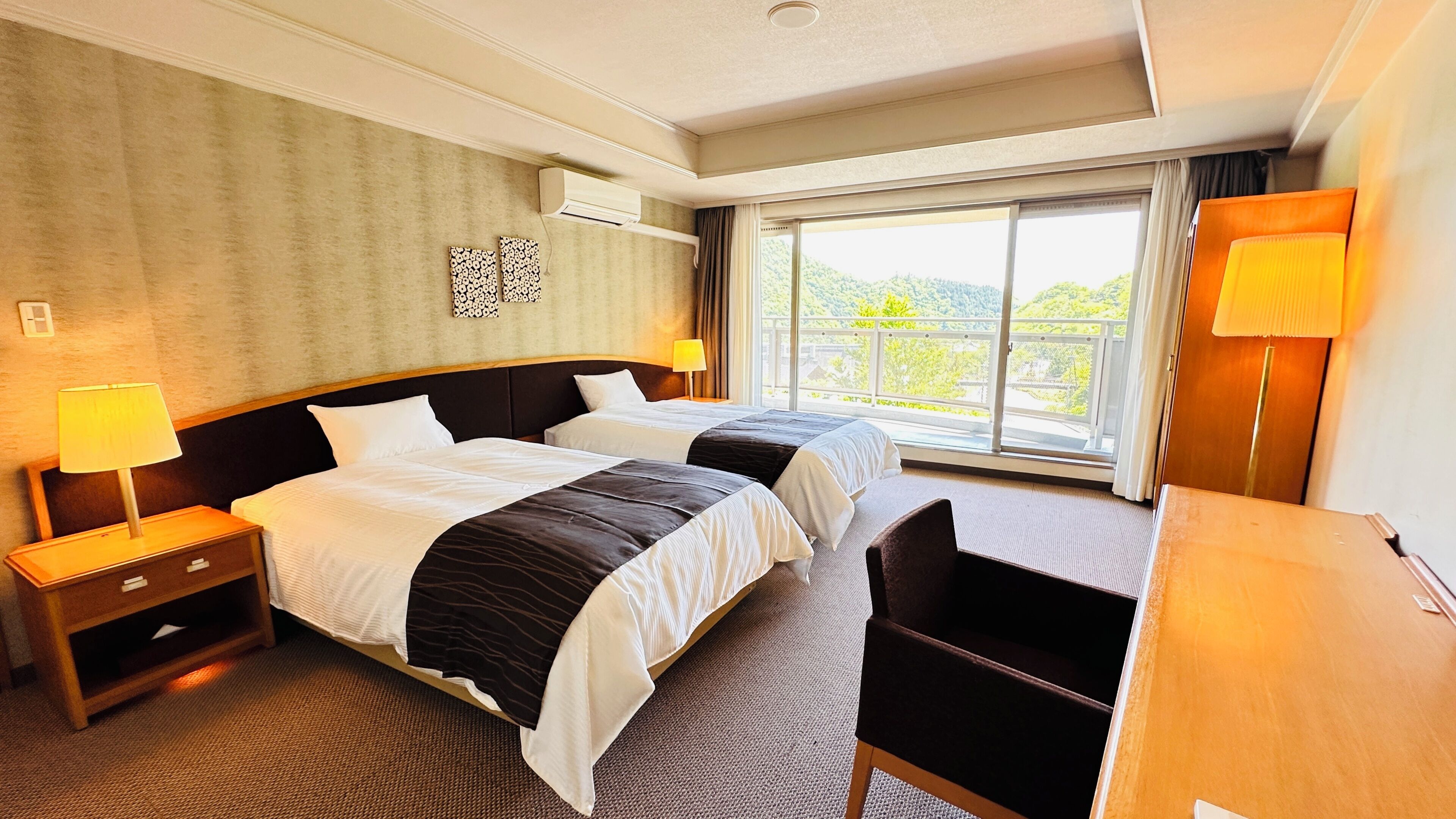 Quarto de hotel com duas camas, uma mesa, uma cadeira, uma luminária e uma janela grande com vista.