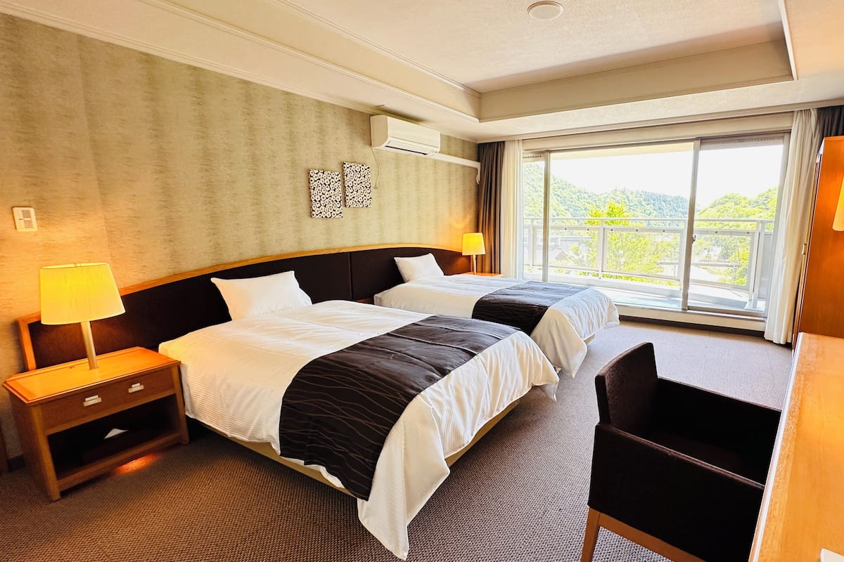 Habitación de hotel con dos camas, un escritorio, una silla, una lámpara y un ventanal con vistas.