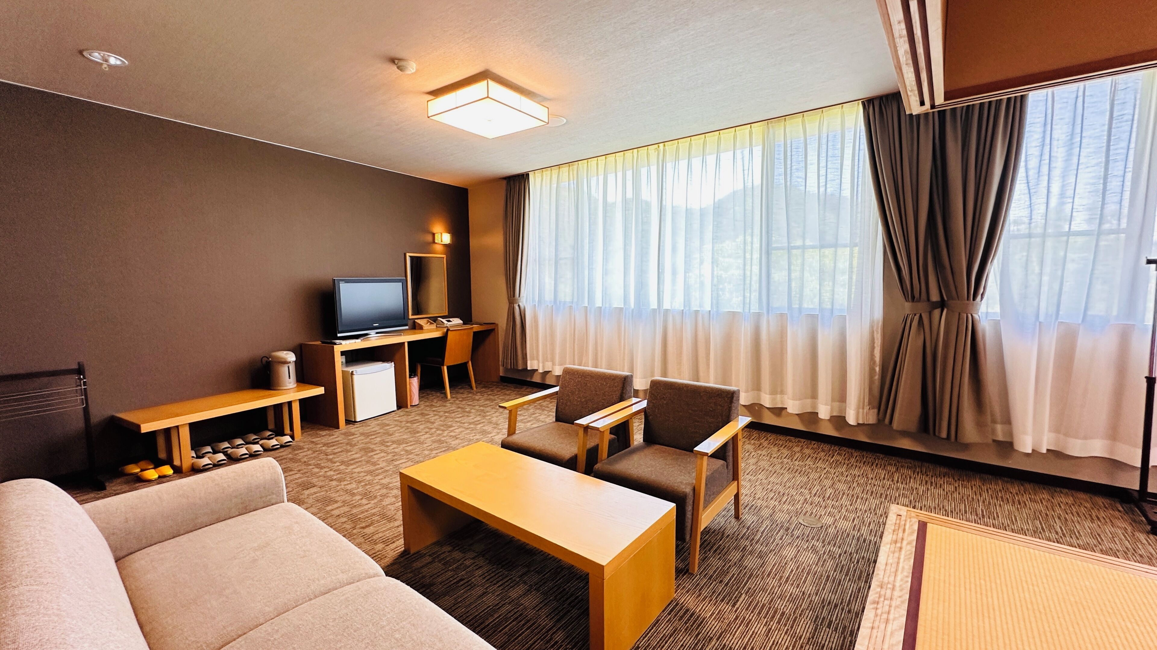 Quarto de hotel com sofá, poltronas, televisão, escrivaninha e uma janela grande com cortinas.
