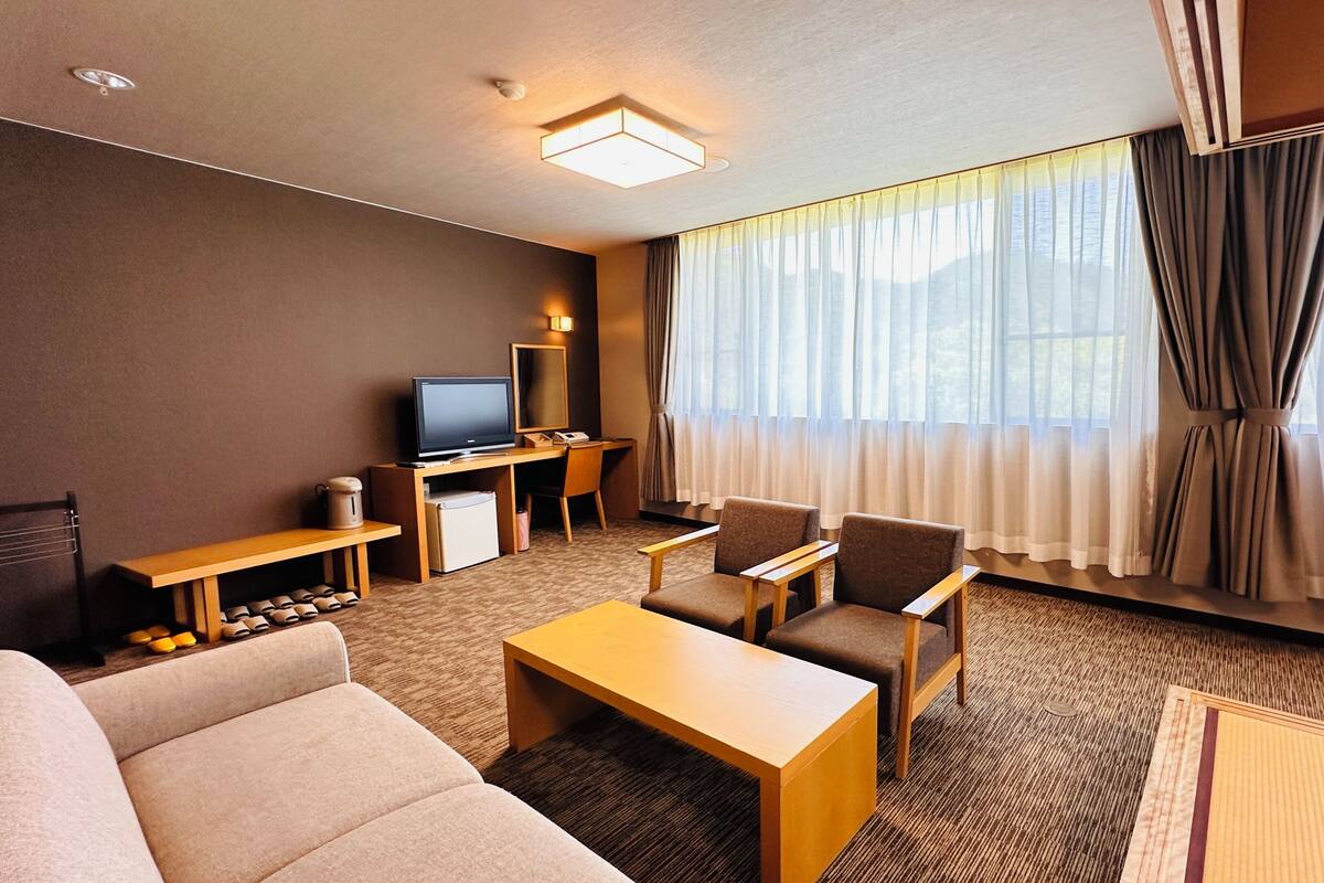 Habitación de hotel con sofá, sillones, televisión, escritorio y un amplio ventanal con cortinas.