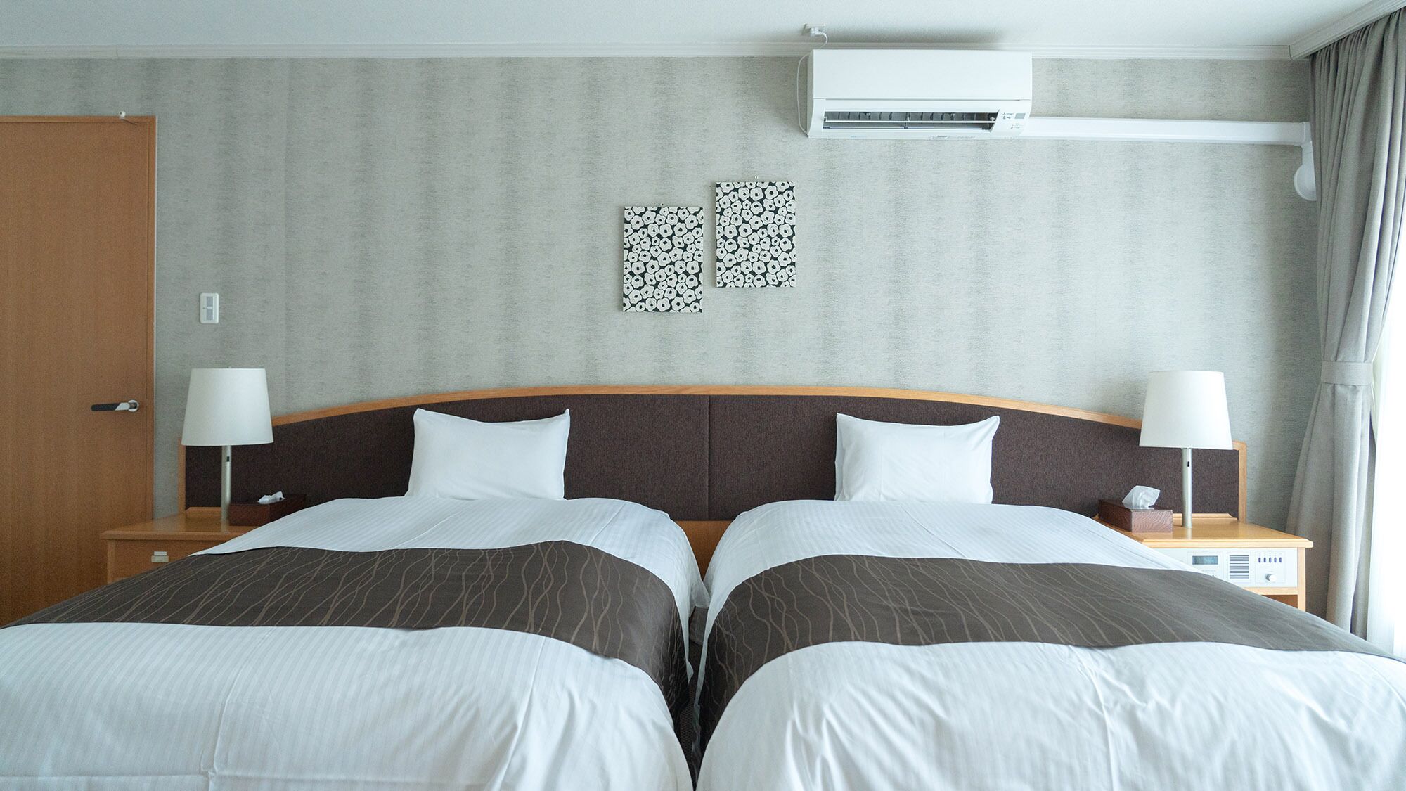 Una habitación de hotel con dos camas, un cabecero de madera, un aire acondicionado instalado en la pared y una mesita de noche con lámpara.