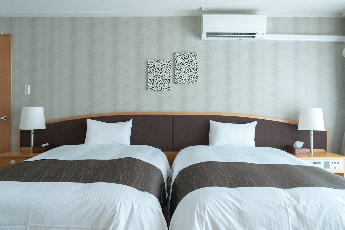 Una habitación de hotel con dos camas, un cabecero de madera, un aire acondicionado instalado en la pared y una mesita de noche con lámpara.