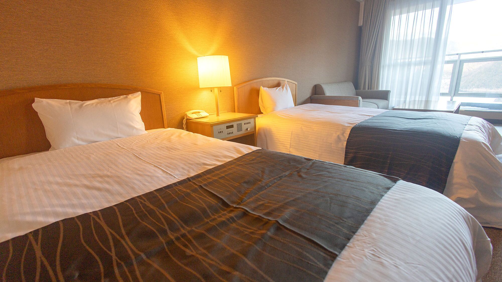 Habitación de hotel con dos camas, una mesita de noche con lámpara, una silla y una ventana con cortinas.