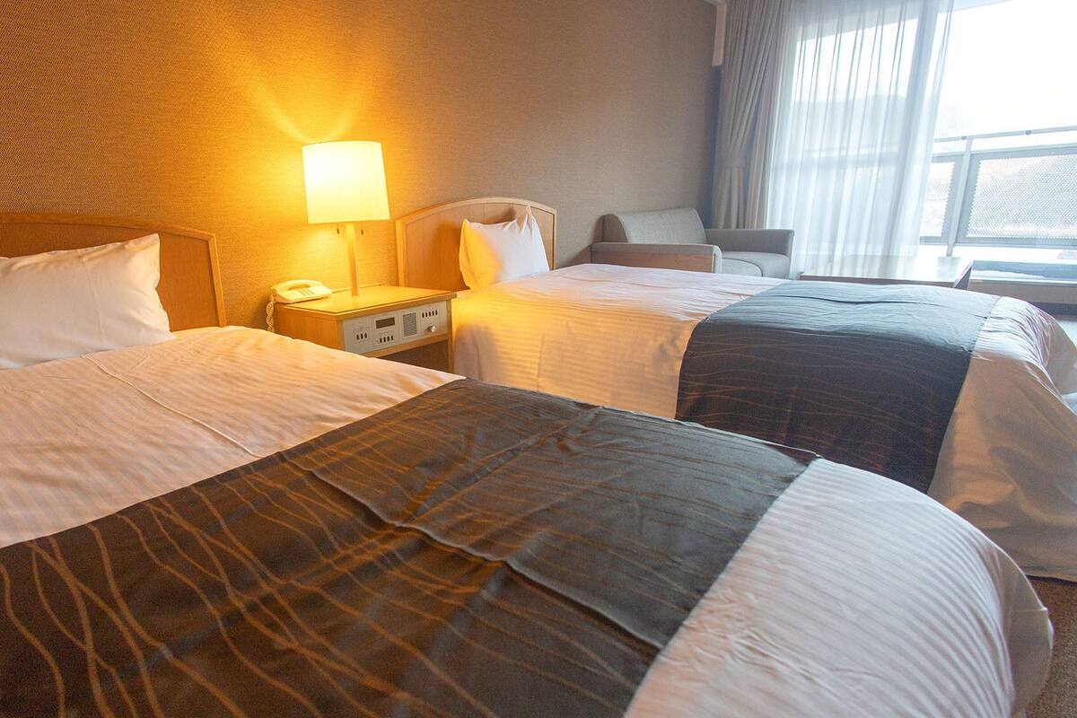 Habitación de hotel con dos camas, una mesita de noche con lámpara, una silla y una ventana con cortinas.