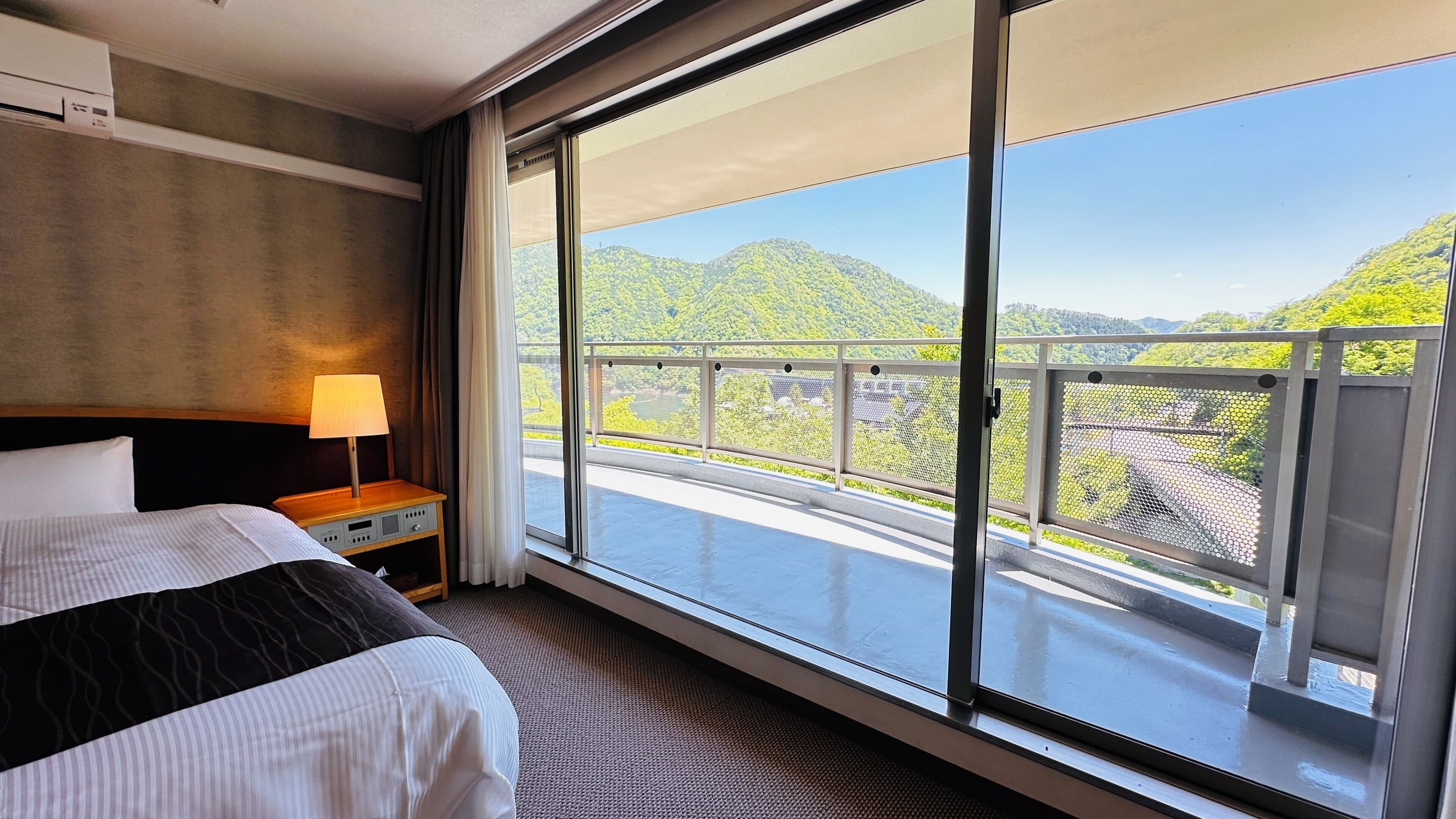 Quarto de hotel com uma janela grande, uma cama, uma mesinha de cabeceira e um abajur.