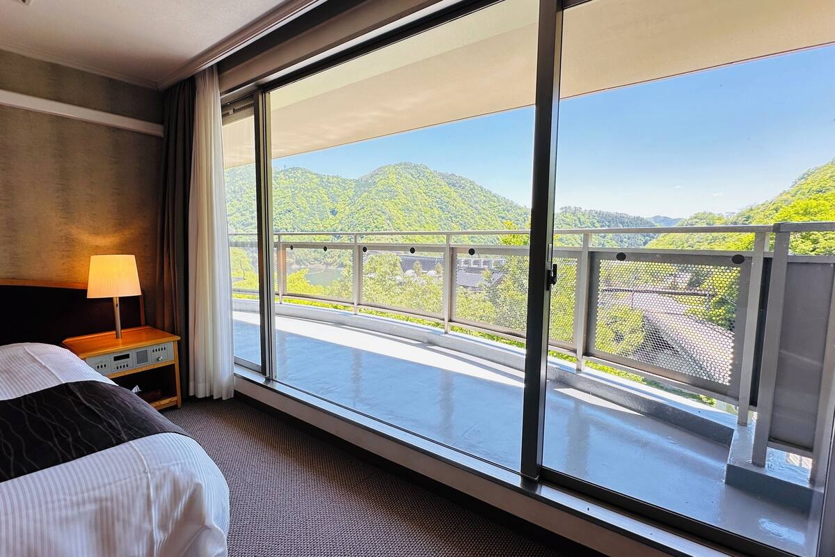 Una habitación de hotel con un ventanal grande, una cama, una mesita de noche y una lámpara.