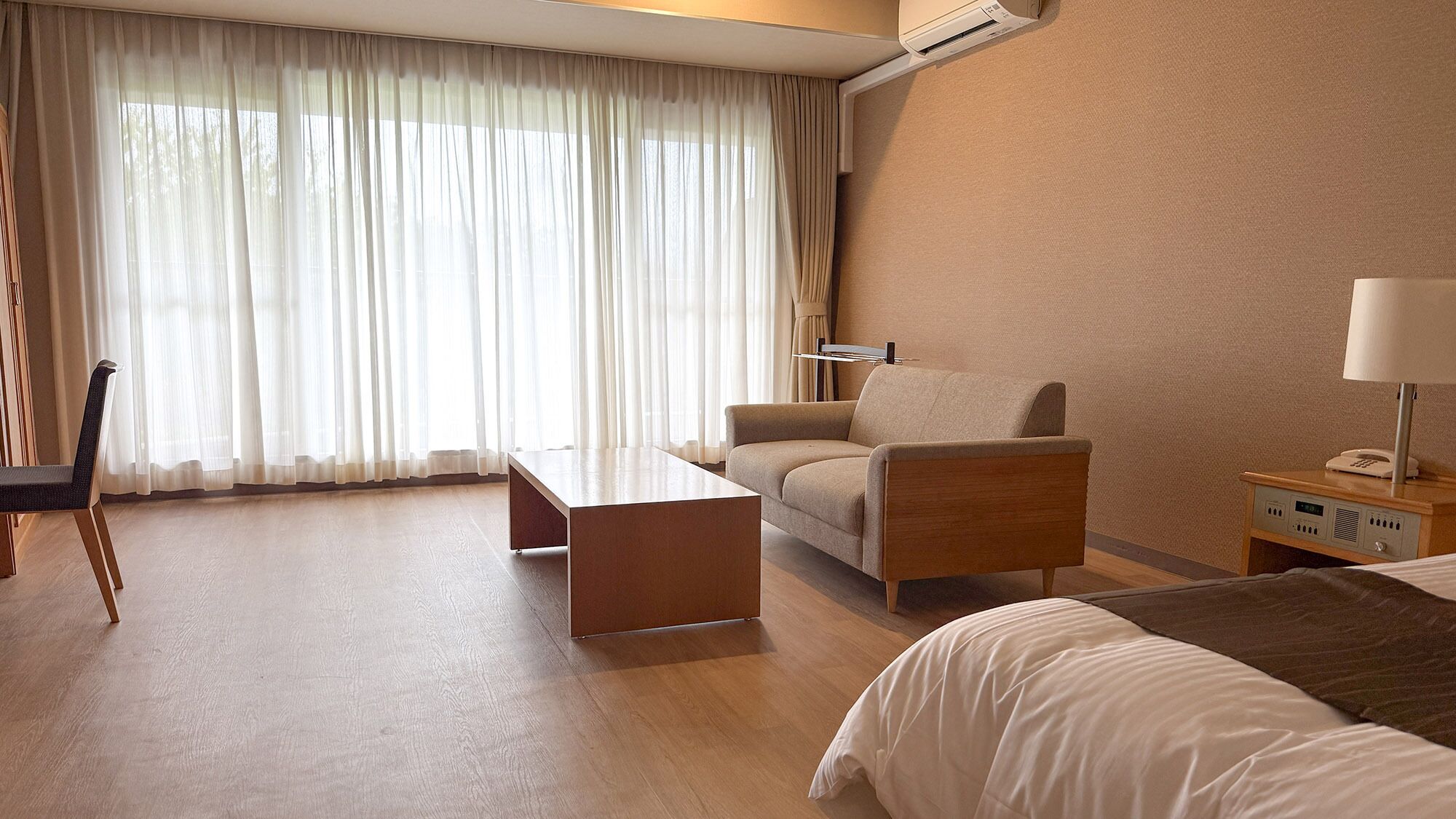 Una habitación de hotel con una cama, un sofá, una mesa, una silla, una mesita de noche con lámpara y una ventana con cortinas.