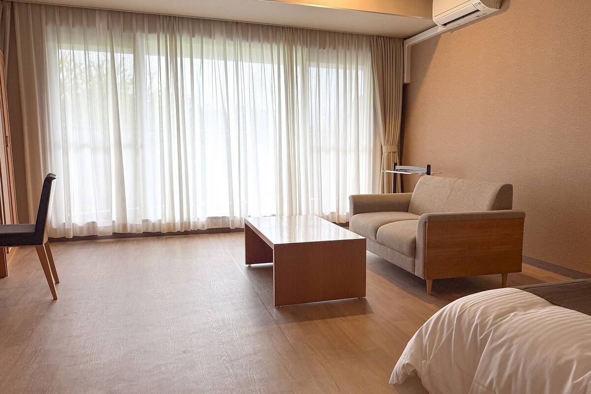 Una habitación de hotel con una cama, un sofá, una mesa, una silla, una mesita de noche con lámpara y una ventana con cortinas.