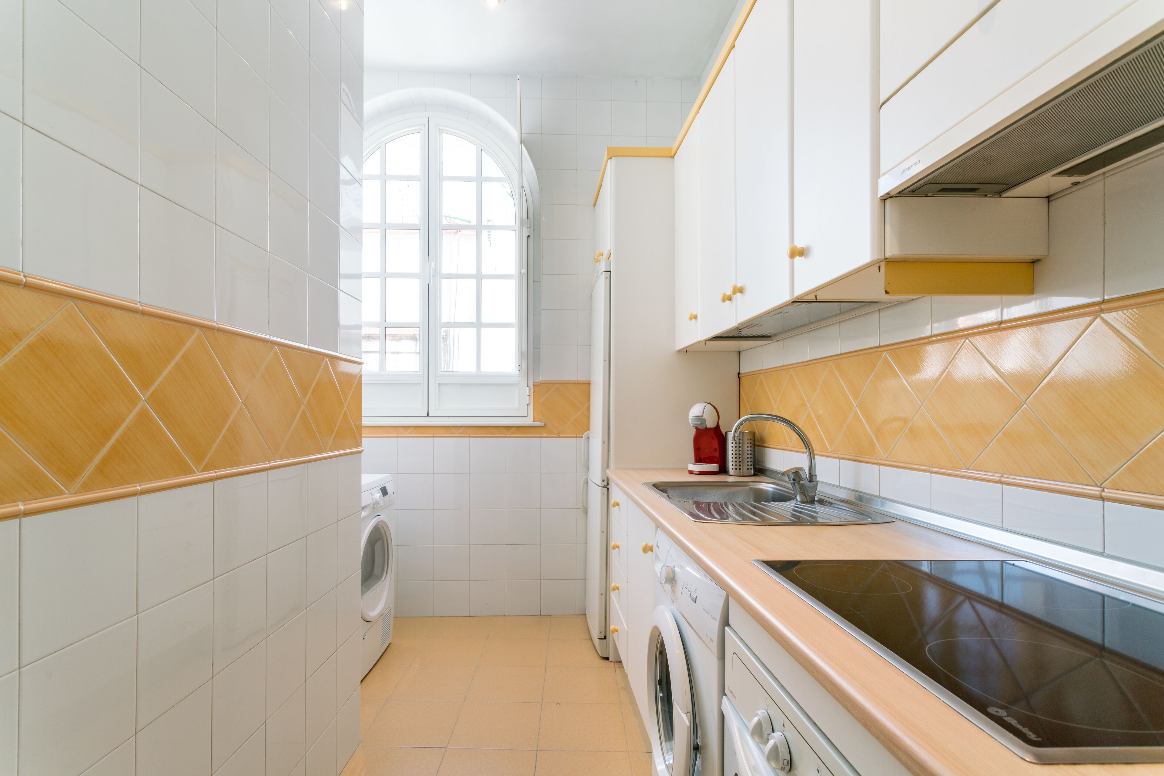 A compact kitchen with white and beige tiles, a built-in washing machine, a sink, and a stove.