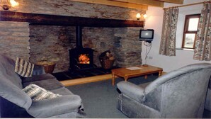 A cozy living room with a stone fireplace, a television, and two grey armchairs. - Pine Cottage - Sheeps Head Peninsula (Bantry)