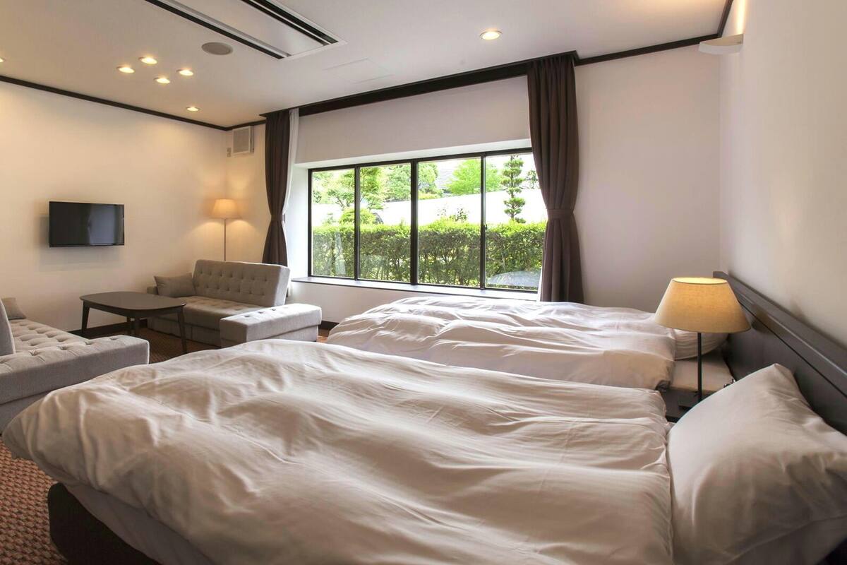 Ein Hotelzimmer mit zwei Betten, einem Schlafsofa, einem kleinen Tisch, einem Fernseher und einem Fenster mit Vorhängen.