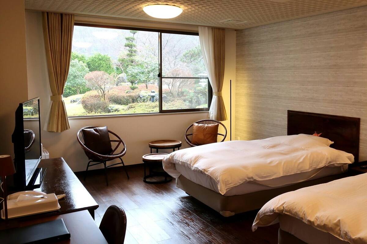 Ein Hotelzimmer mit zwei Betten, einem Schreibtisch mit einem Laptop, einem Sessel und Blick auf einen Garten durch ein großes Fenster.