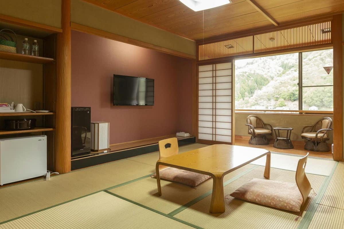 Ein traditioneller japanischer Raum mit Tatami-Böden, einem niedrigen Tisch, Stühlen, einem Fernseher und Blick auf Kirschblüten.