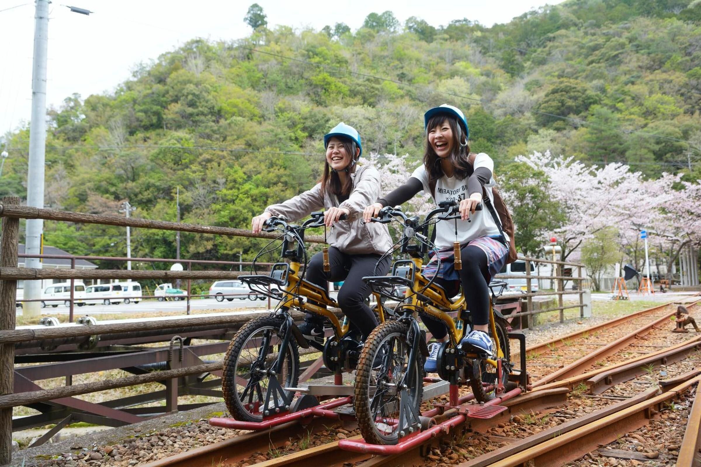 Dos personas andan en bicicleta sobre vías de tren, con una colina boscosa al fondo.