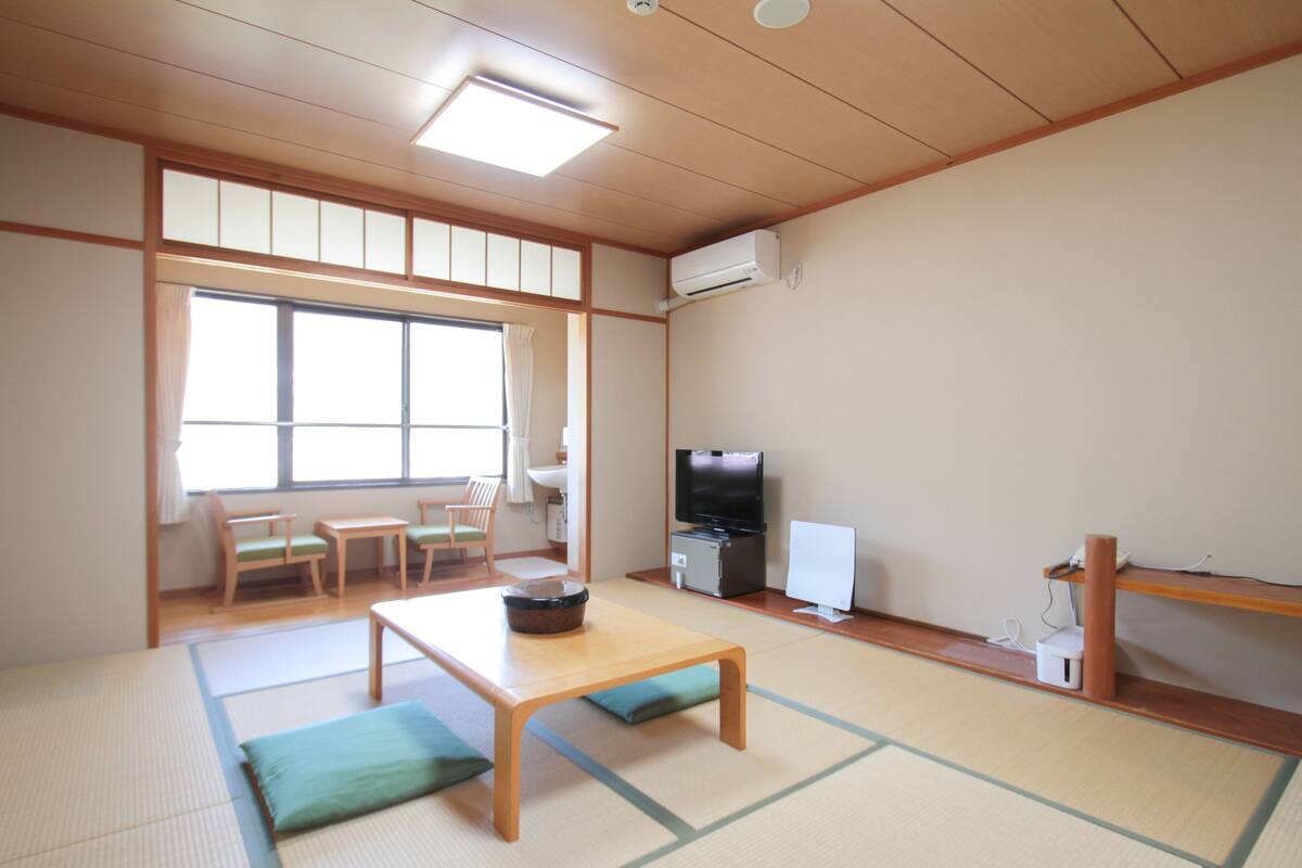 Ein traditioneller japanischer Raum mit Tatami-Böden, einem niedrigen Tisch mit Kissen, einem Fernseher und einem kleinen Kühlschrank.