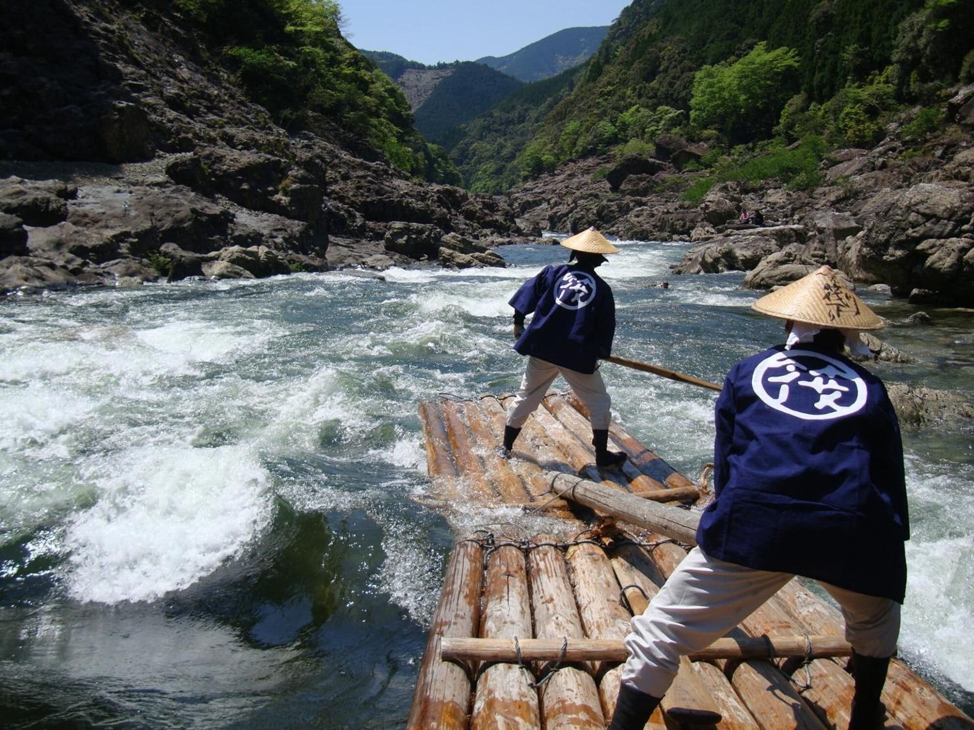 Two individuals on a bamboo raft navigating a river surrounded by rocky terrain and green hills.