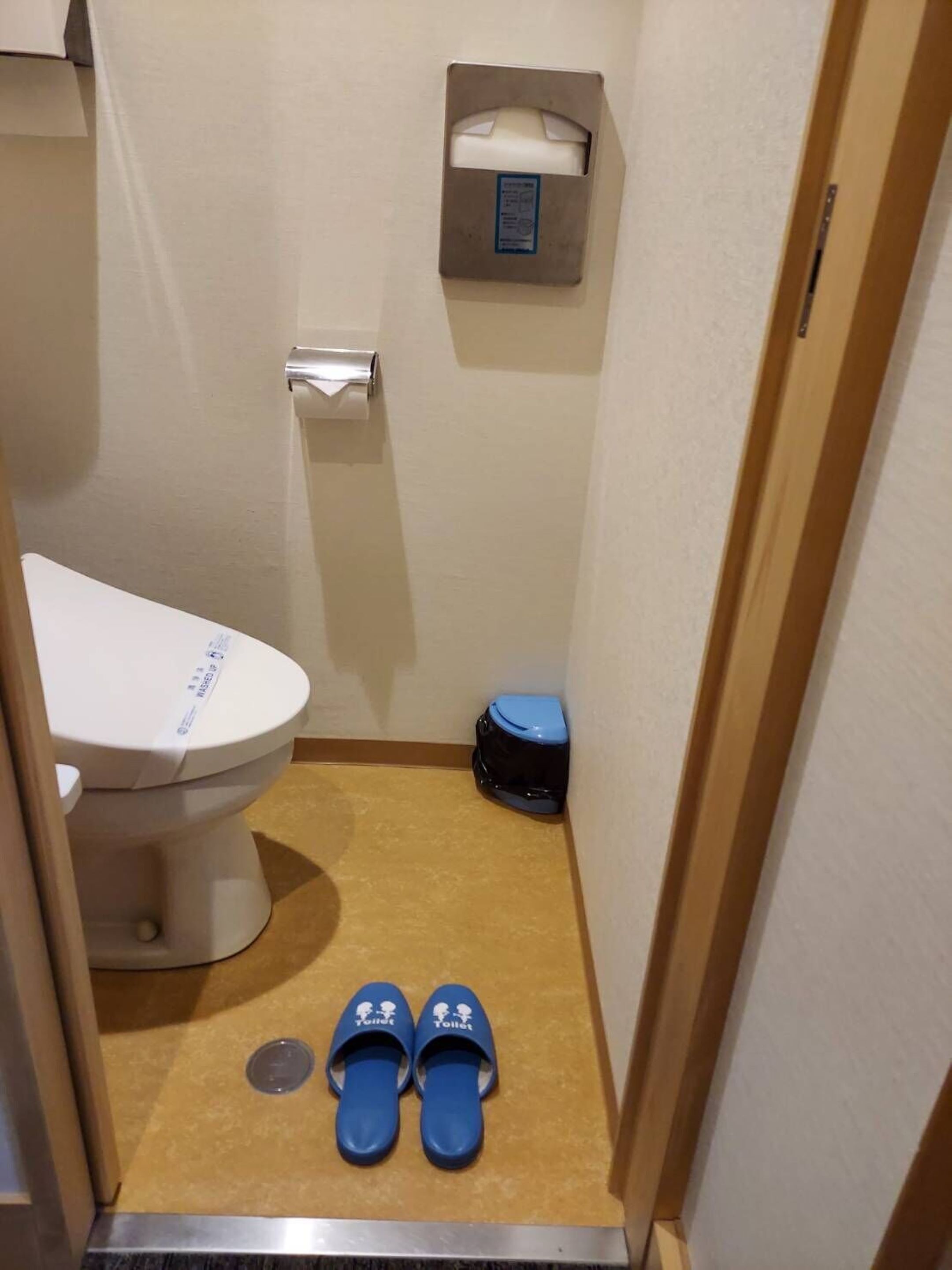 Un baño pequeño con un inodoro, un cubo de basura y un par de pantuflas en el suelo.