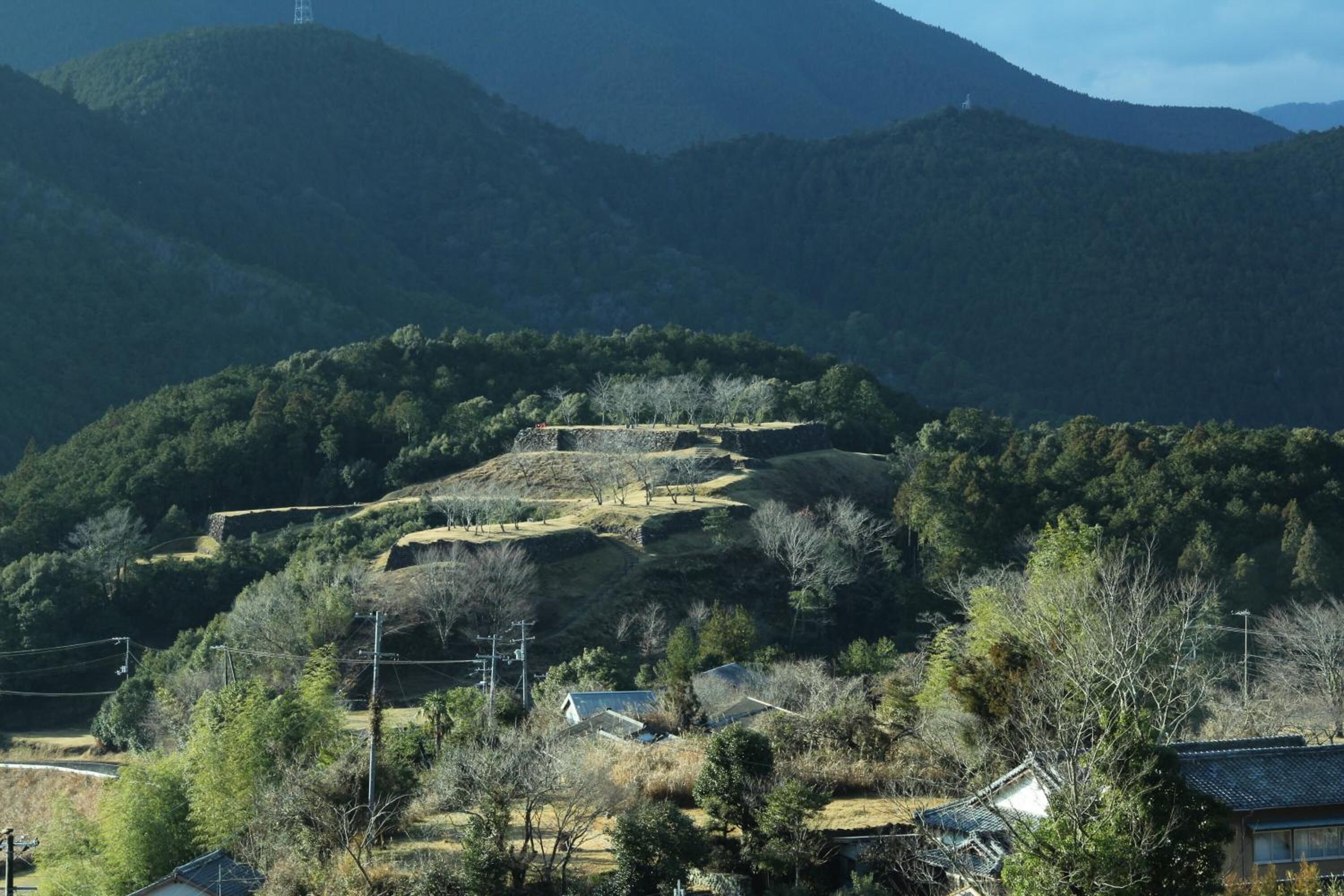 A rural landscape with a traditional Japanese house surrounded by hills and sparse vegetation.