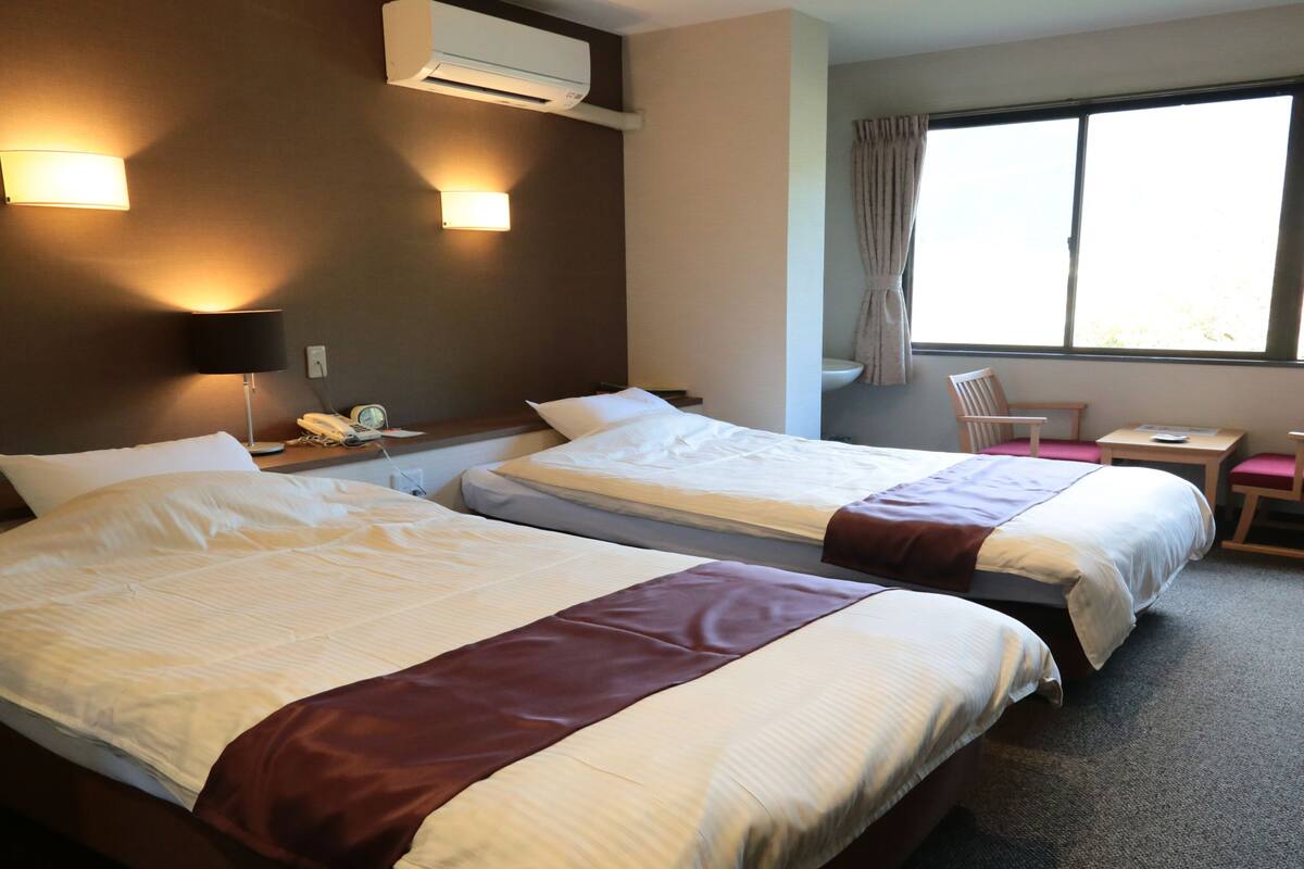 Ein Hotelzimmer mit zwei Betten, einem Schreibtisch, einem Stuhl, einem Fenster und einer Klimaanlage.