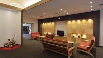 A modern lobby with a wooden reception desk, red cushioned seating, a flat-screen TV, and a decorative plant.