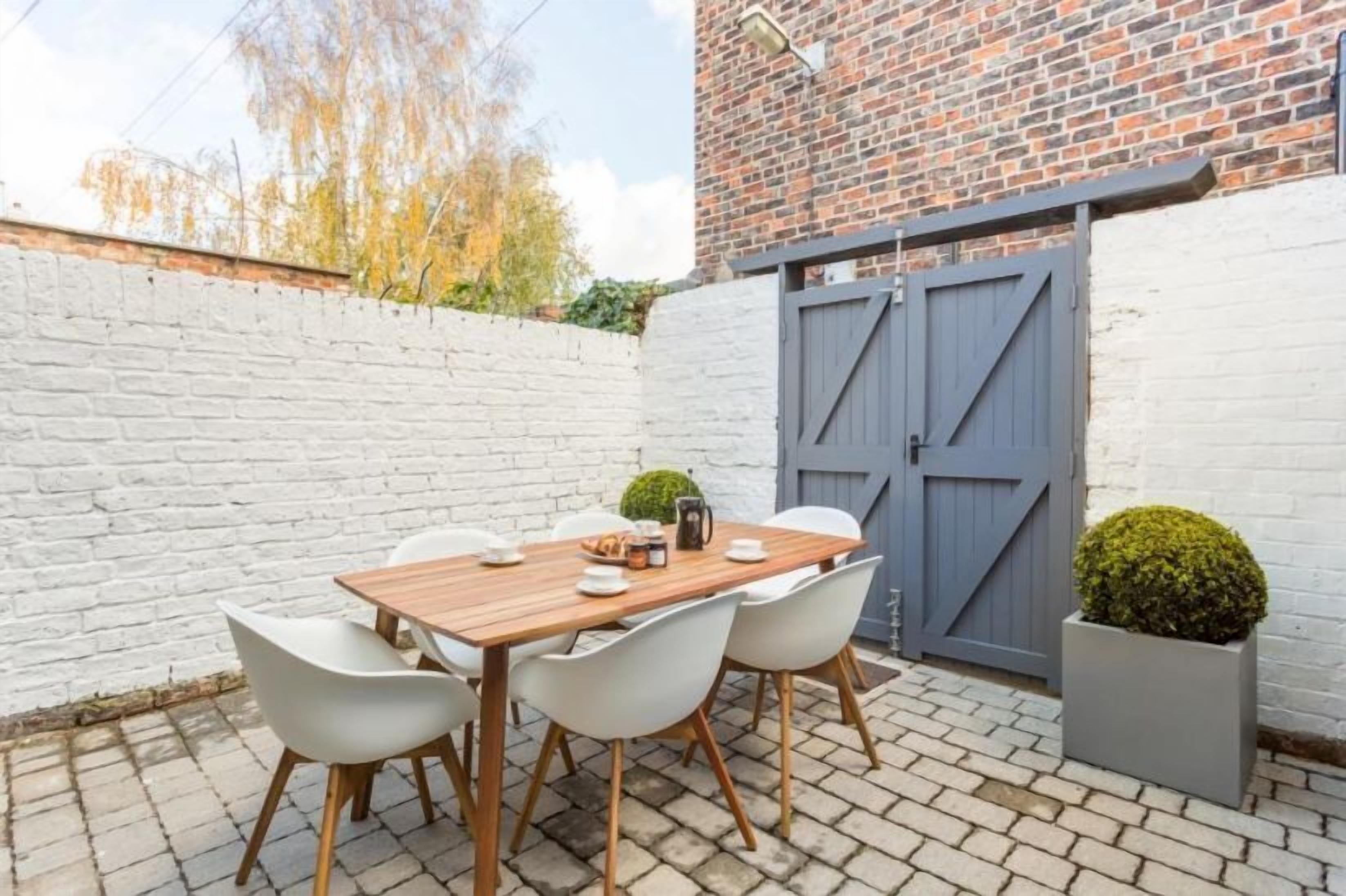 Een terras met een houten tafel en witte stoelen, een bakstenen muur en een grijze poort.