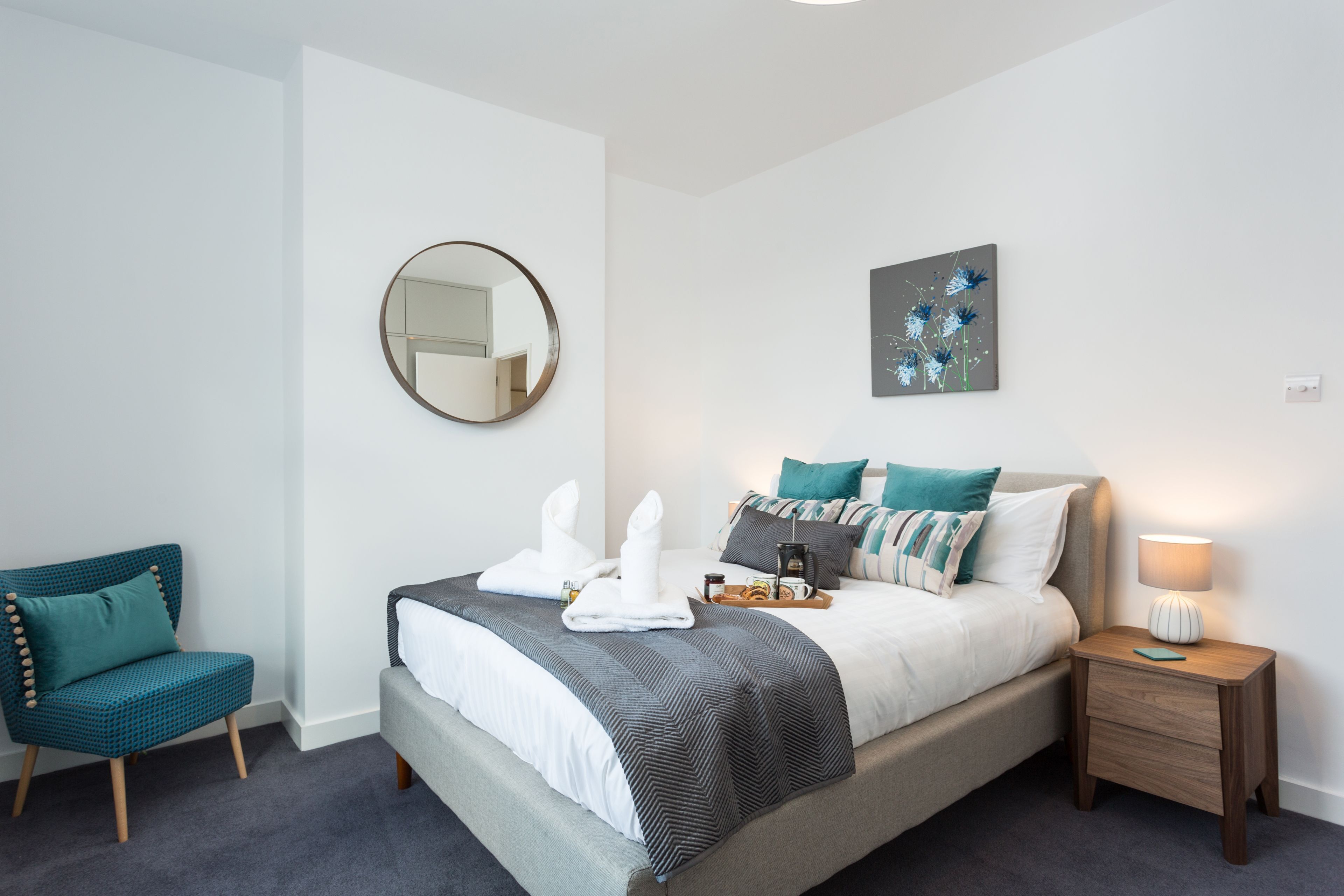 A neatly arranged bedroom with a bed, bedside table, chair, and artwork on the wall.