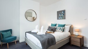 Un dormitorio ordenado con cama, mesita de noche, silla y una obra de arte en la pared.