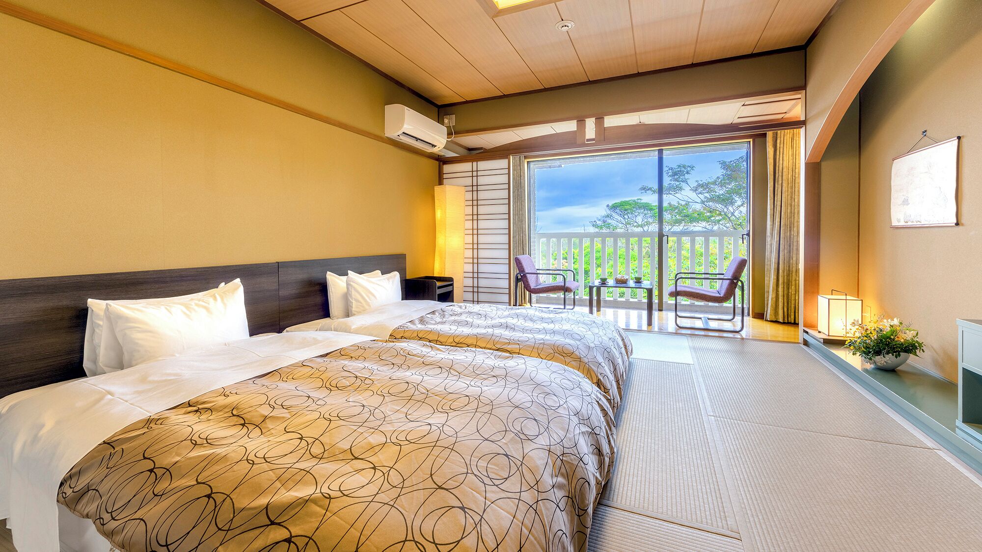 A hotel room with a large bed, a balcony with chairs, and a view of greenery.