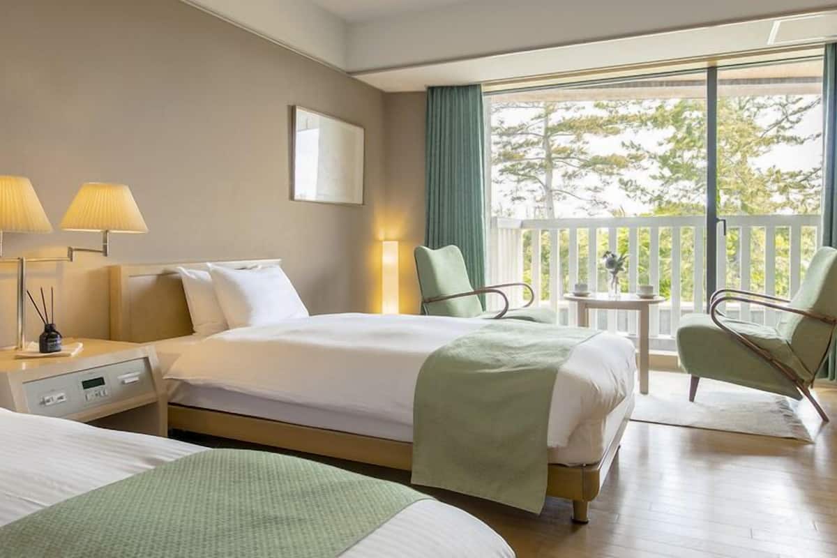 Habitación de hotel con dos camas, una mesita de noche, una lámpara, una silla y un balcón con vistas.
